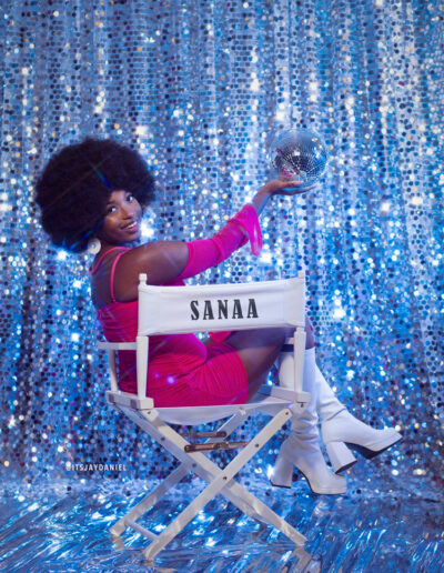 High school senior with afro hair. The student stands in front of a colorful, retro-inspired backdrop featuring a geometric pattern and vibrant shades of blue, reds, and pins. The student's outfit features a pink dress, and knee high white gogo boots, which are reminiscent of 70s fashion. The student looks confident and proud, with a big smile on their face.