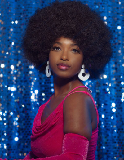 High school senior with afro hair. The student stands in front of a colorful, retro-inspired backdrop featuring a geometric pattern and vibrant shades of blue, reds, and pins. The student's outfit features a pink dress, and knee high white gogo boots, which are reminiscent of 70s fashion. The student looks confident and proud, with a big smile on their face.