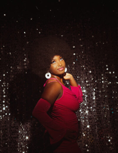 High school senior with afro hair. The student stands in front of a colorful, retro-inspired backdrop featuring a geometric pattern and vibrant shades of blue, reds, and pins. The student's outfit features a pink dress, and knee high white gogo boots, which are reminiscent of 70s fashion. The student looks confident and proud, with a big smile on their face.